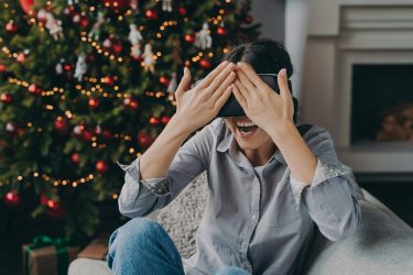 Joyful young woman in VR headset glasses sitting on cozy sofa next to decorated Xmas tree at home